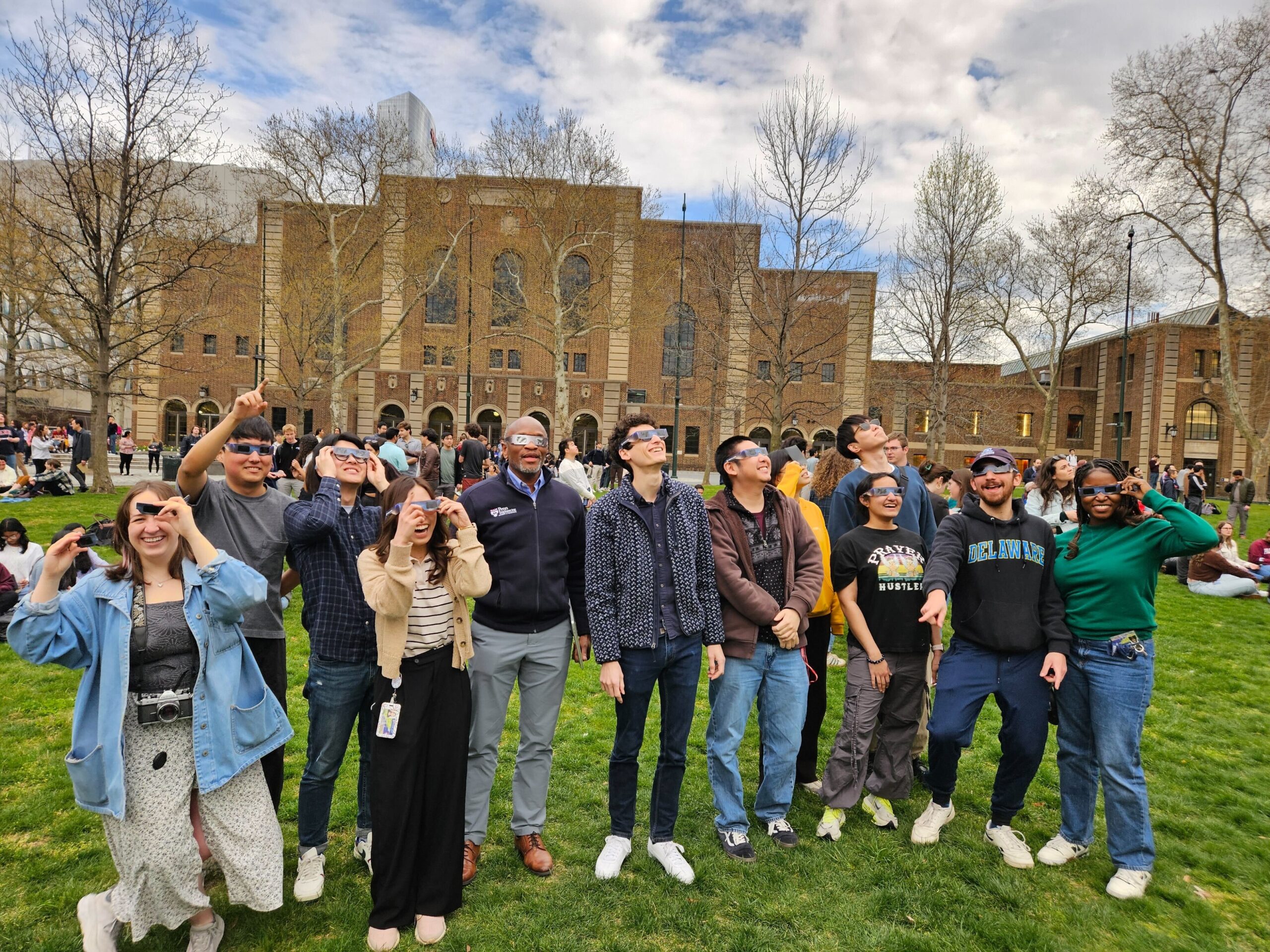 a group of college students and faculty wearing solar eclipse glasses look at the sky from a lawn on a college campus