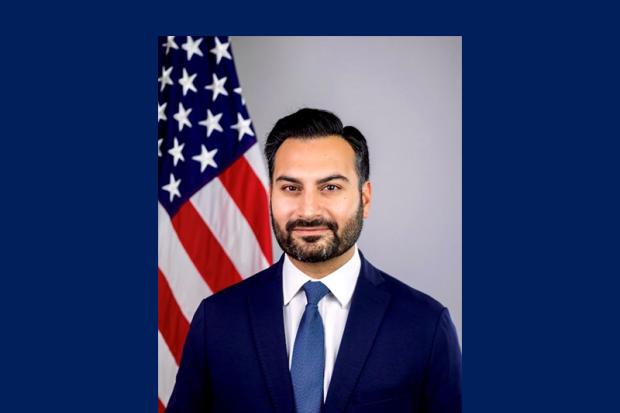 man with dark hair in blue suit in front of US flag