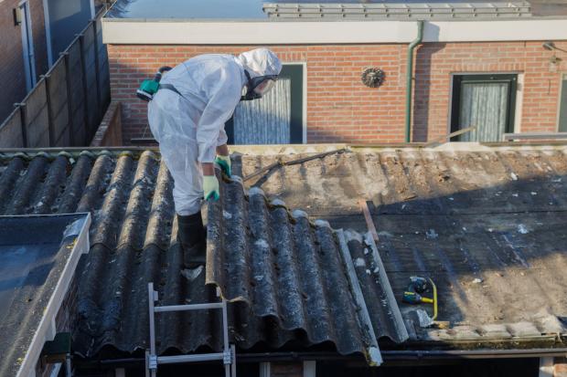A person in a hazmat suit removes a piece of roofing from a roof.