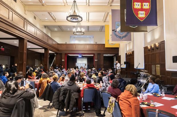Students sit at tables to eat breakfast in a large room with wooden panelling, chandeliers, and university flags. 